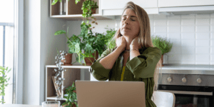 Woman sitting at her kitchen table with her eyes closed and hands on her neck, experiencing fatigue and discomfort associated with high ferritin levels symptoms.