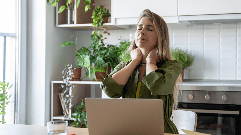 Woman sitting at her kitchen table with her eyes closed and hands on her neck, experiencing fatigue and discomfort associated with high ferritin levels symptoms.