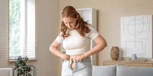 Young woman struggling to button her jeans at home, reflecting unexplained weight changes and bloating associated with hormonal birth control.
