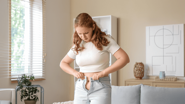 Young woman struggling to button her jeans at home, reflecting unexplained weight changes and bloating associated with hormonal birth control.